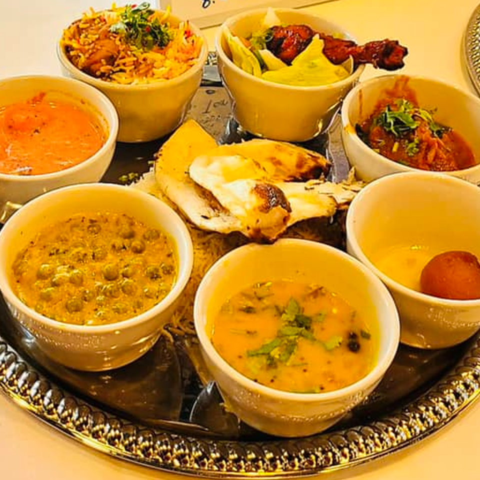 A silver platter holds small bowls of Indian dishes including biryani, curries, naan, and gulab jamun.